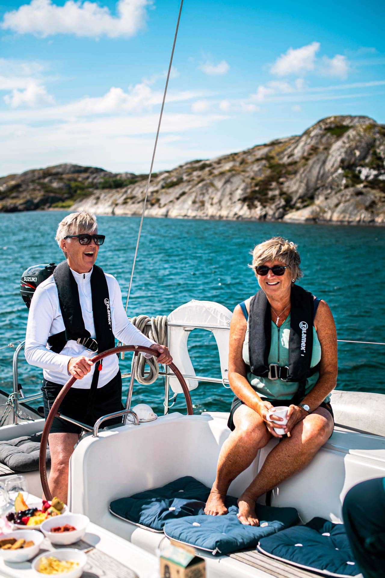 Zwei lachende Frauen an Bord, eine steht am Steuer, die andere trinkt Kaffee.