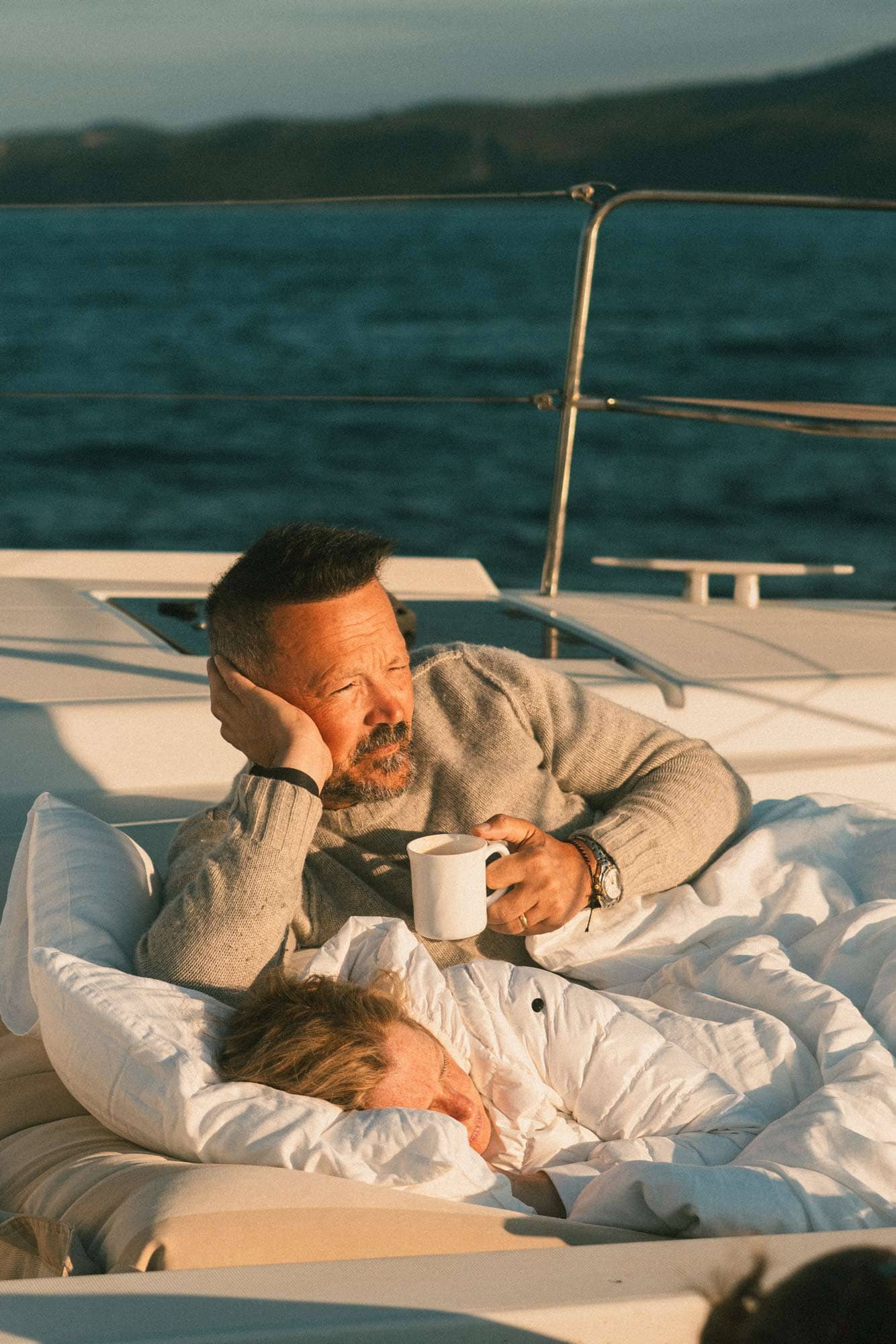 Ein Mann schaut mit einer Tasse Kaffee in der Hand auf einem Segelboot in die Sonne. Eine Frau liegt daneben und schläft.