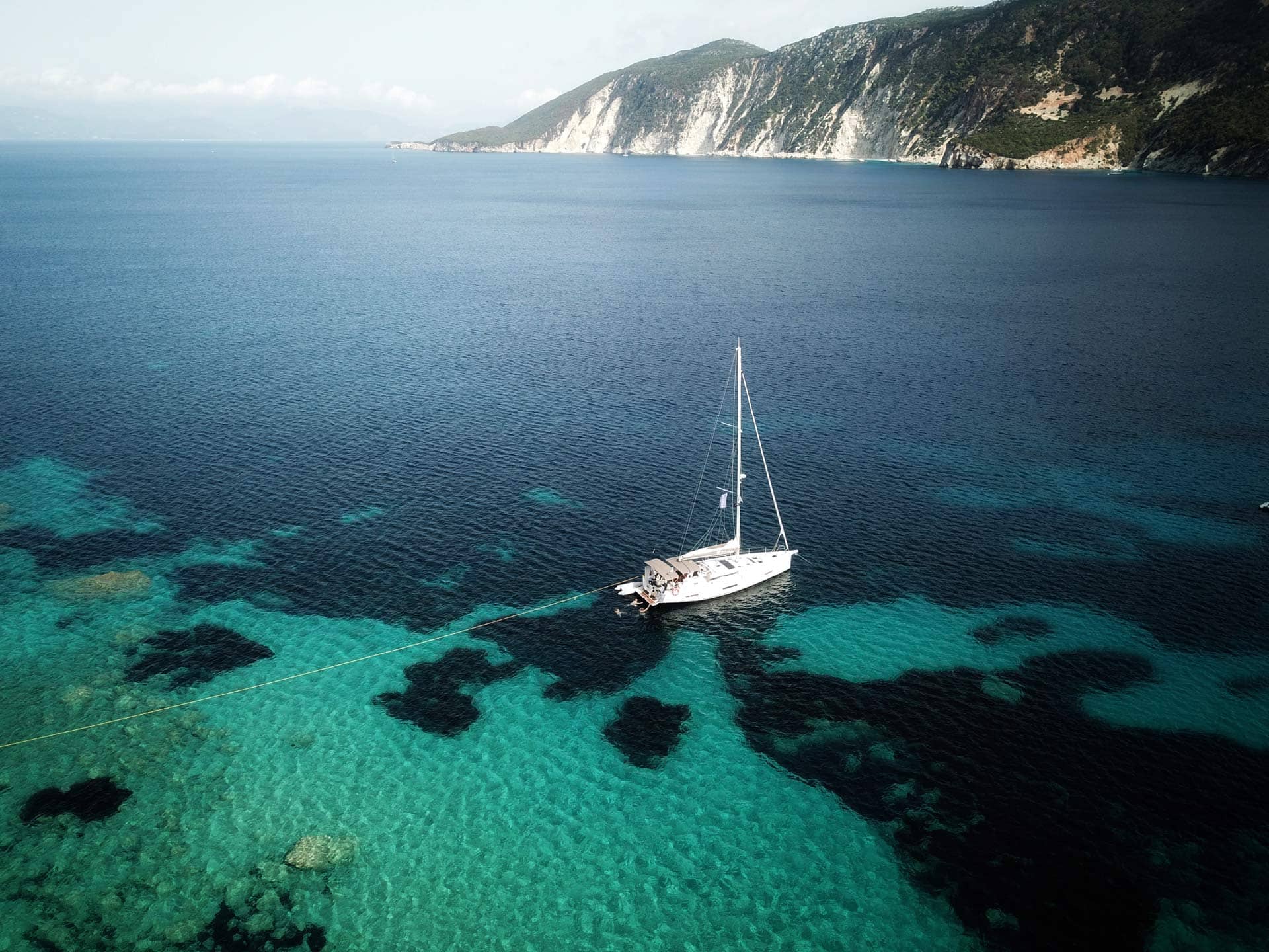 Drohnenaufnahme einer Segelyacht im türkisfarbenen und dunkelblauen Wasser.