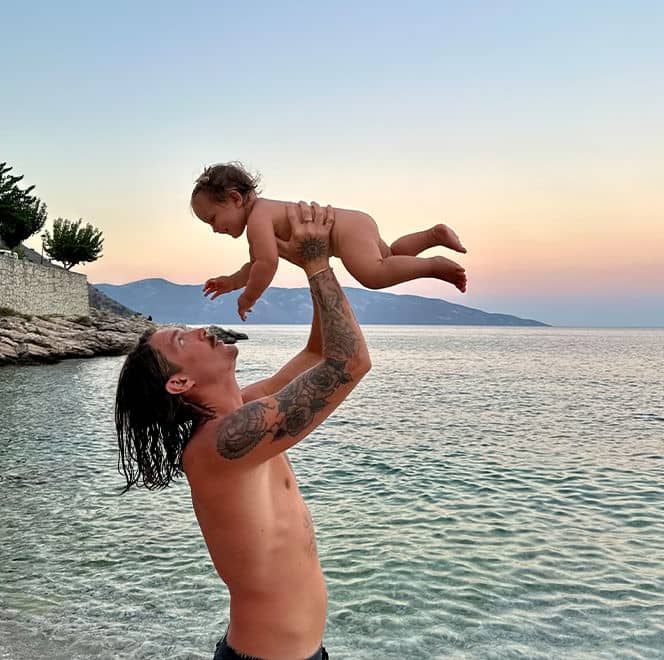 En pappa lyfter upp sitt lilla barn i luften med havet i bakgrunden