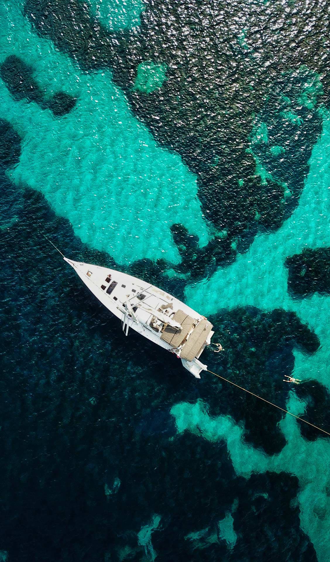 Aerial view of a white monohull in turquoise and dark blue waters.