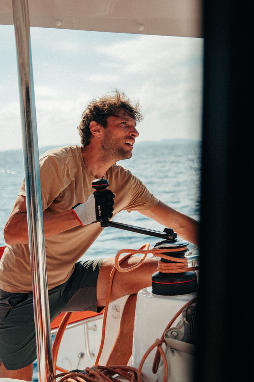 Ein Mann holt an der Winsch eine orangefarbene Leine ein, er trägt einen beigen Pullover und Segelhandschuhe