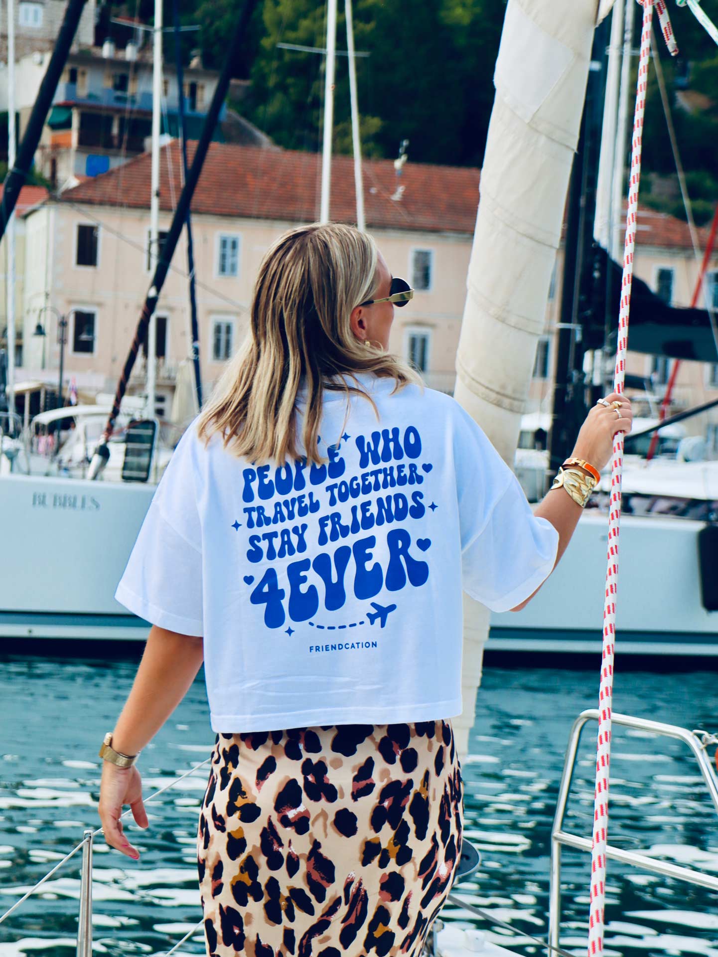 Tjej står på segelbåt och tittar bortåt. Har en tshirt på sig med text. 