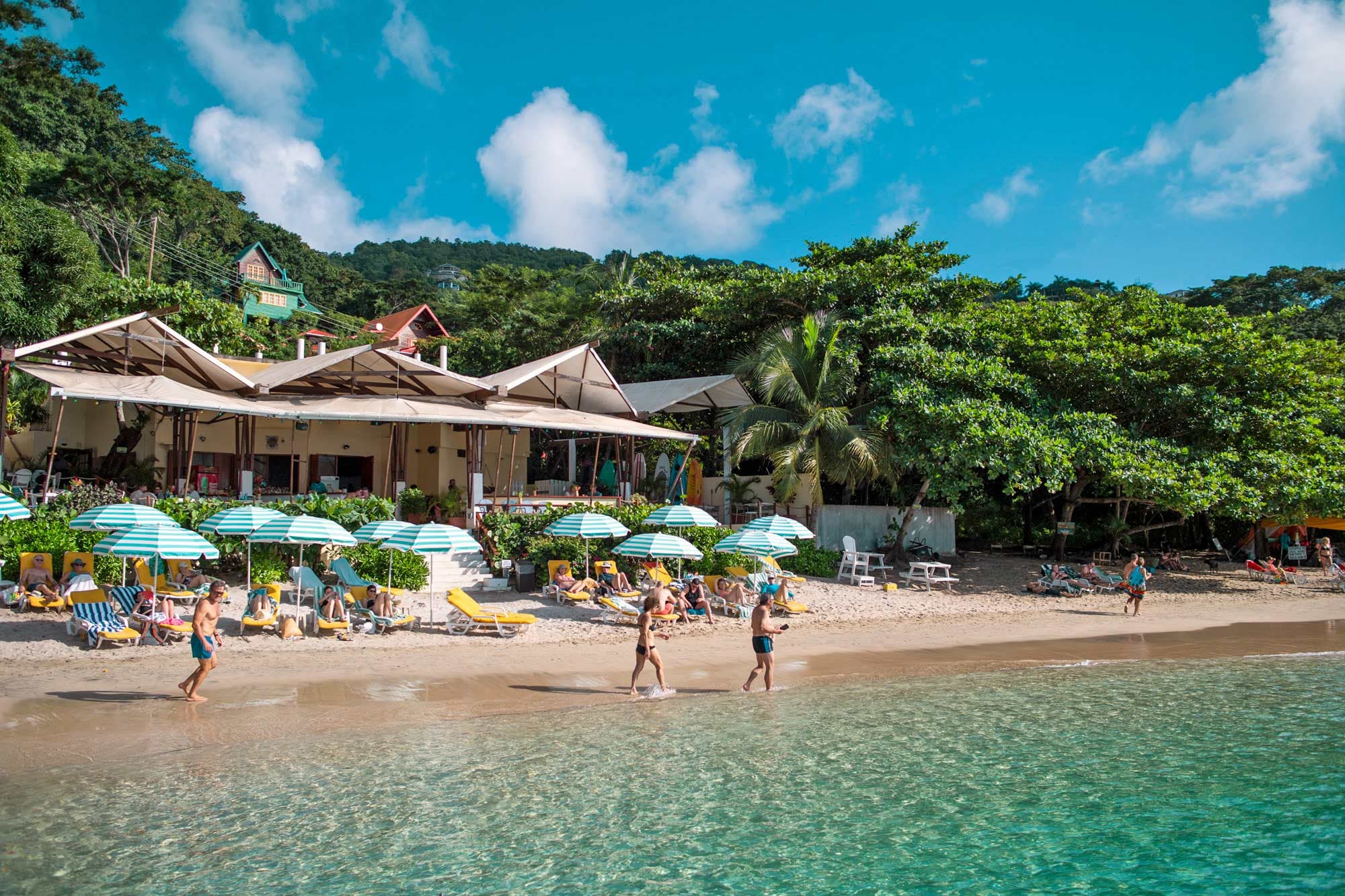Beach in front of a restaurant, 8 striped parasols and a couple walking by the water.