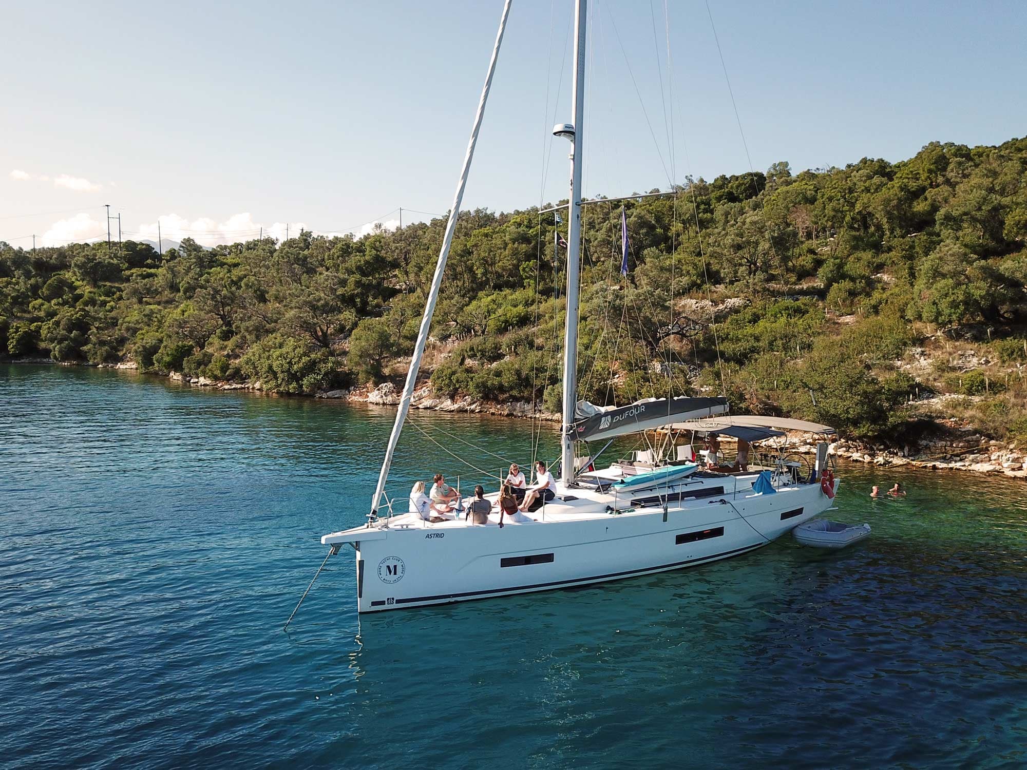A monohull anchored in a bay. A group of people sit on the foredeck in the sun.