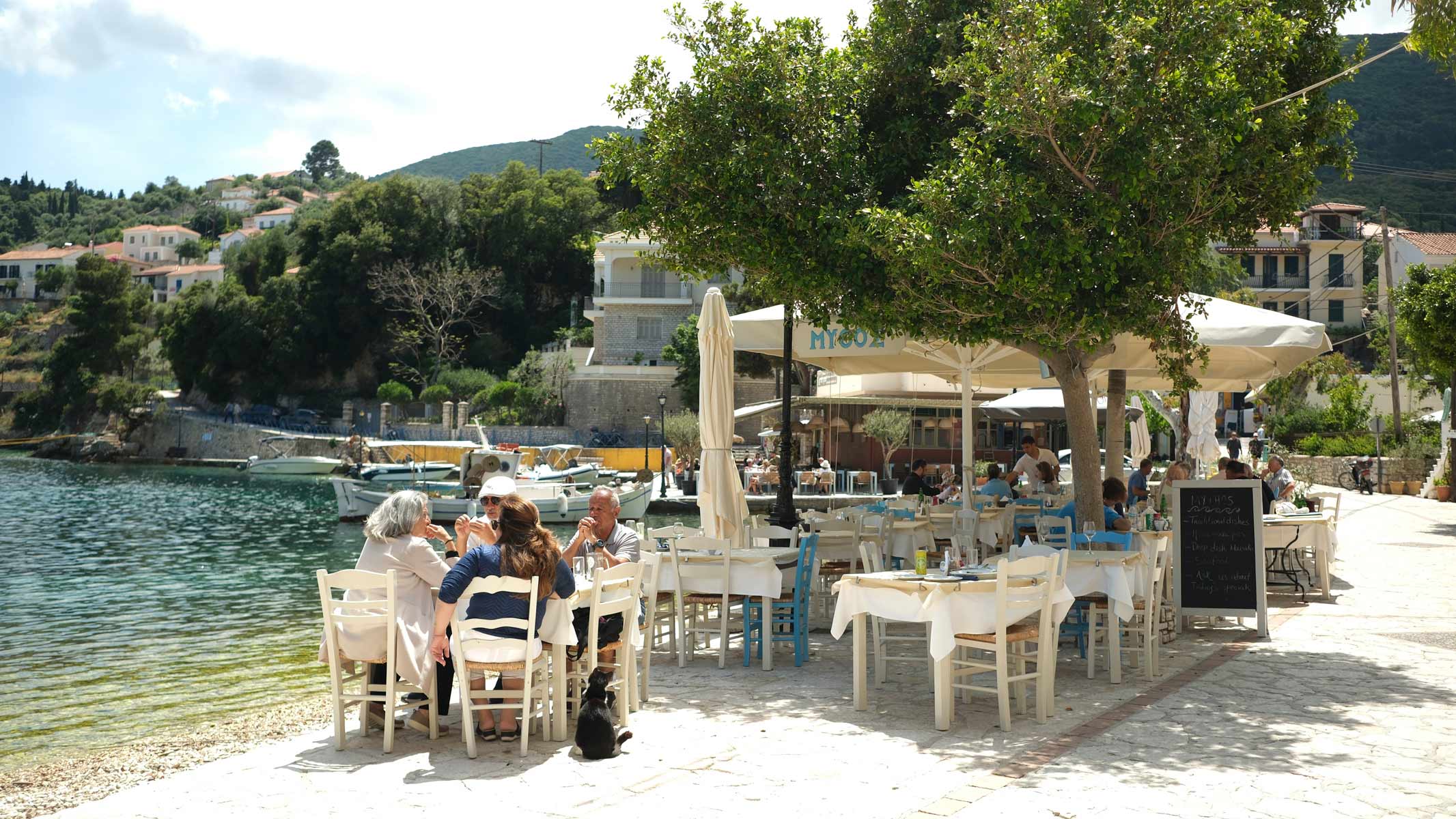 A restaurant with several white tables lining the quay. A group of 4 and a dog sitting at one of the tables.