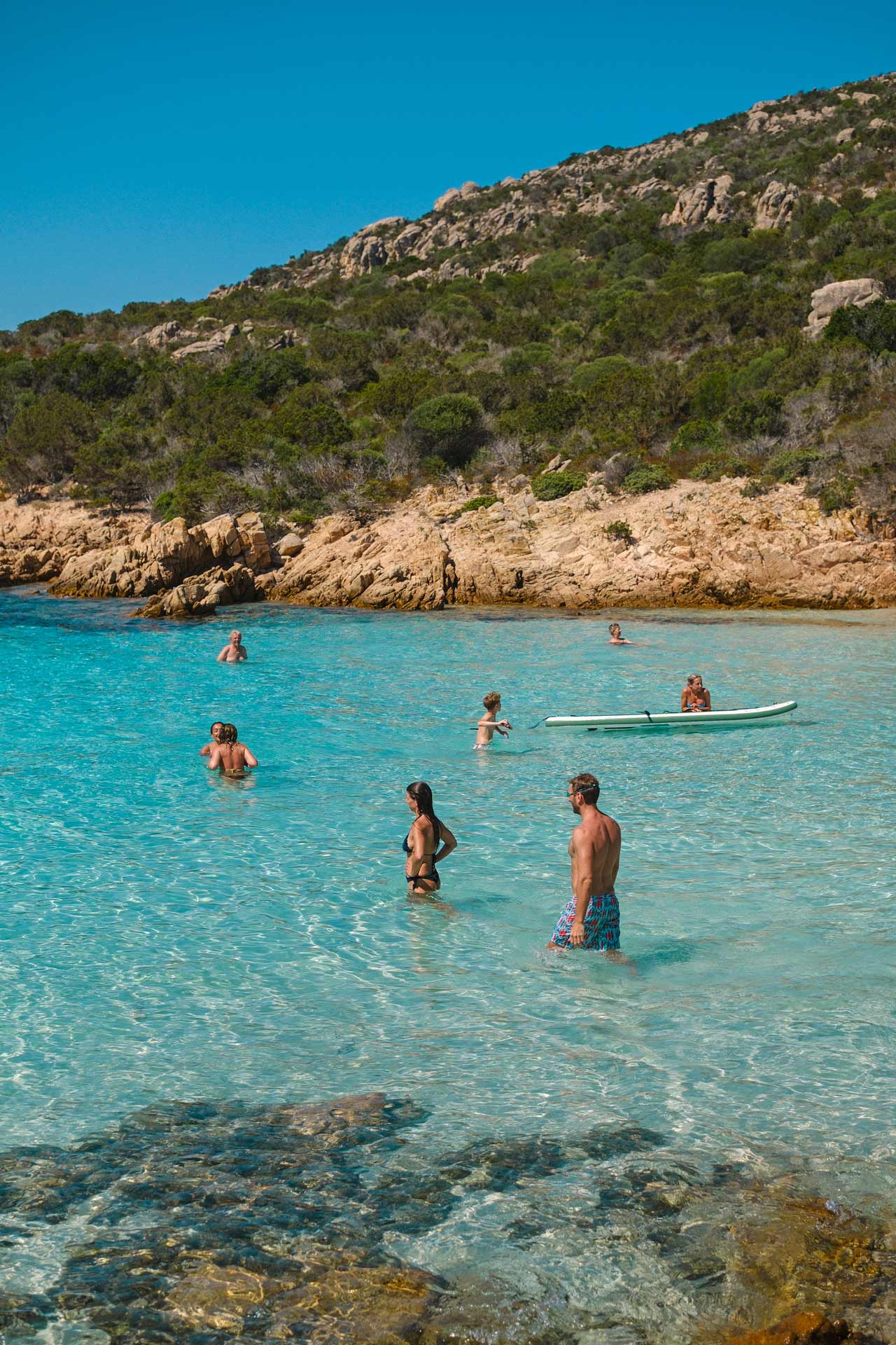 Sky blue bay where guests are swimming; one has a stand up paddle board.