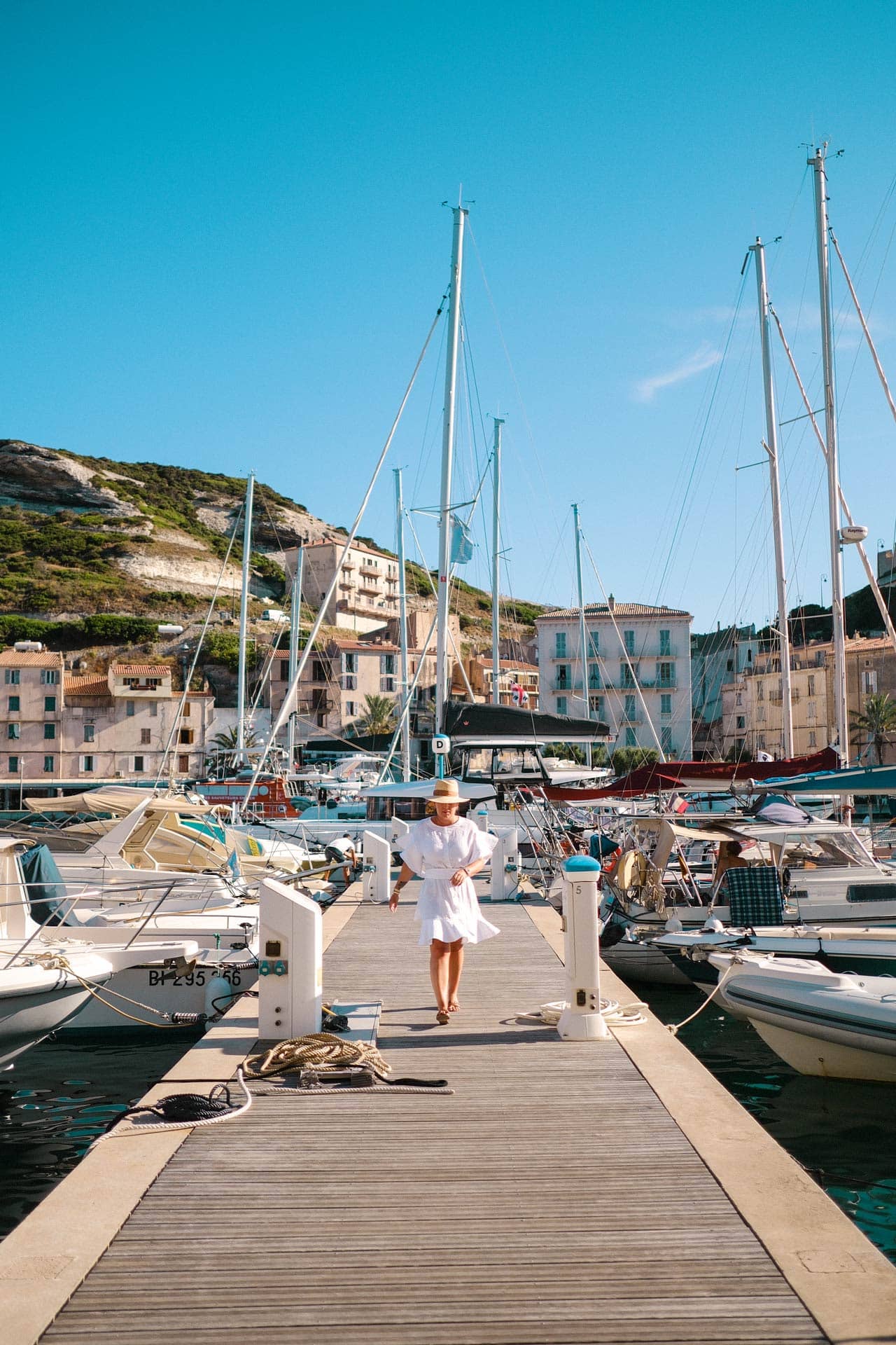 Guest in white dress walking along a jetty lined with boats on either side.