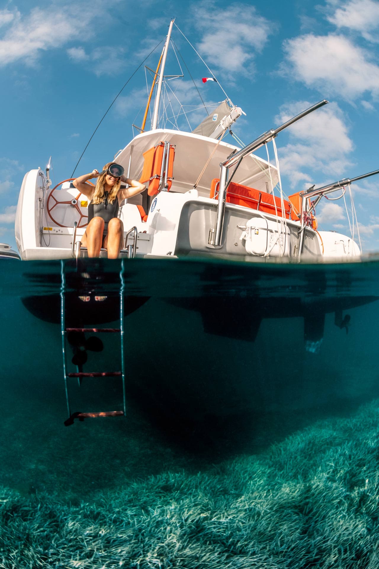 Eine Frau im schwarzen Badeanzug setzt ihre Taucherbrille auf. Halb unter Wasser sieht man Seegras und die Badeleiter