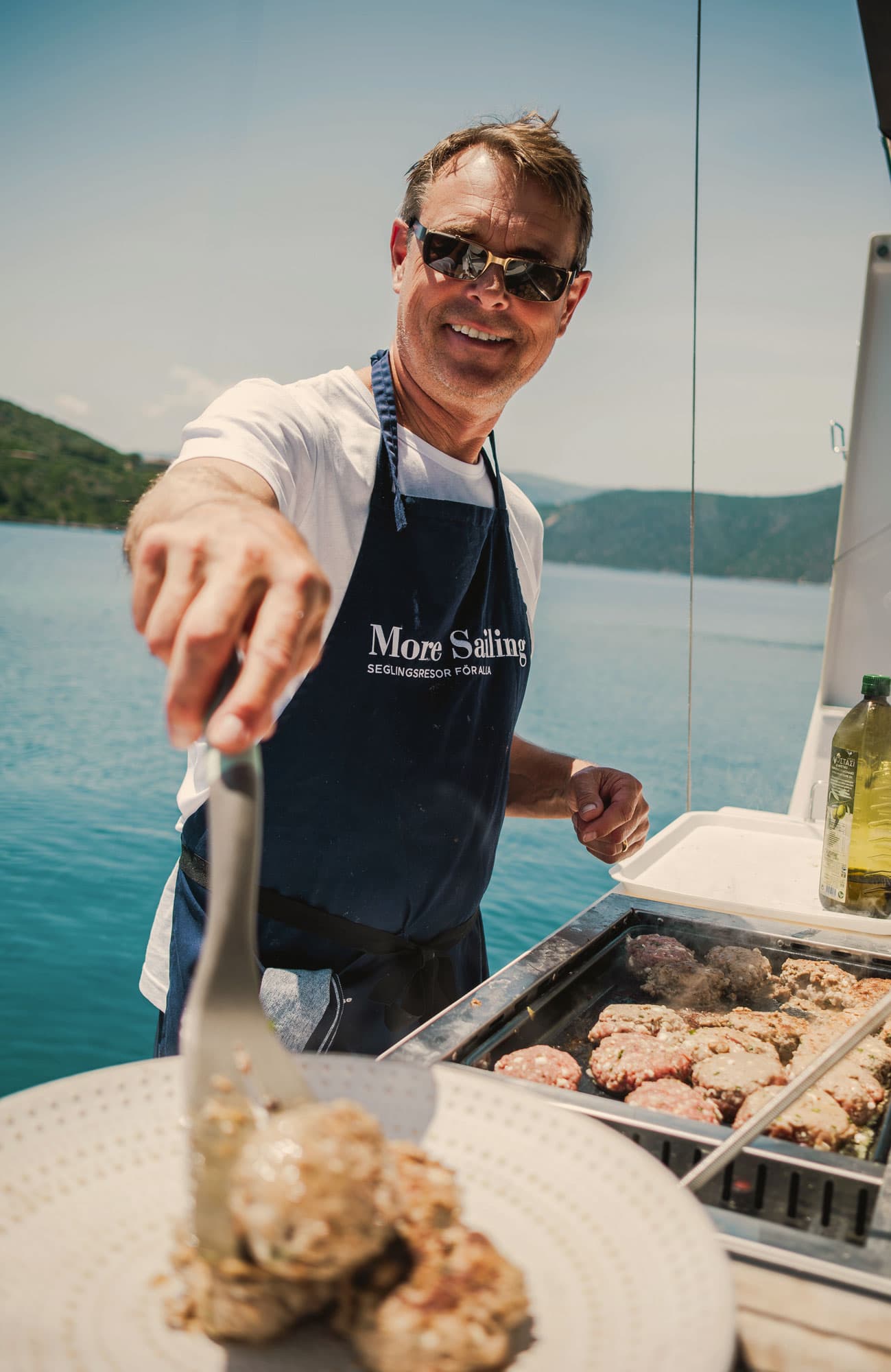Der Gastgeber brät Frikadellen auf der Grillplatte an der Badeplattform des Segelboots