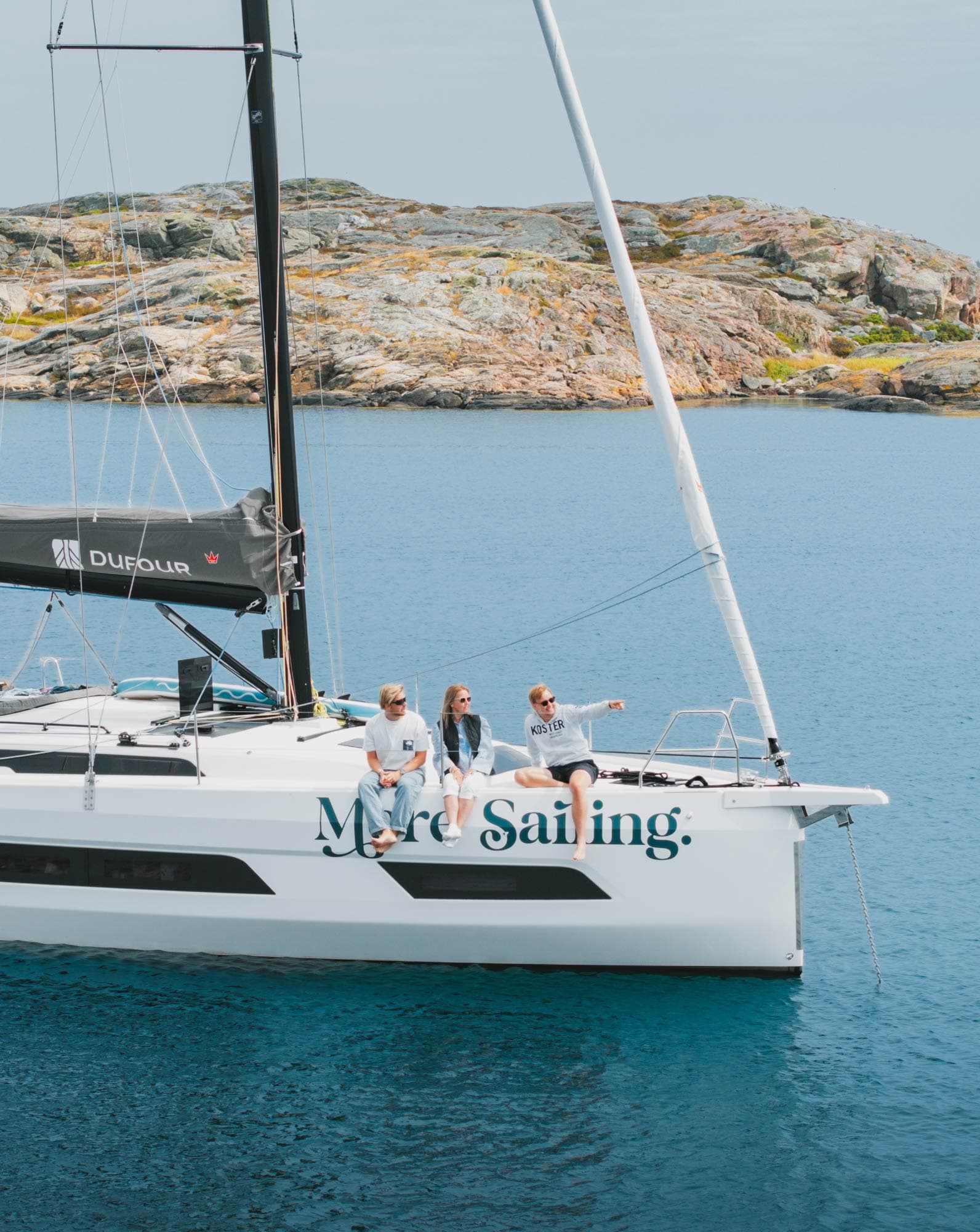 Drei Gäste lassen an der Reling einer More Sailing-Yacht die Beine baumeln.