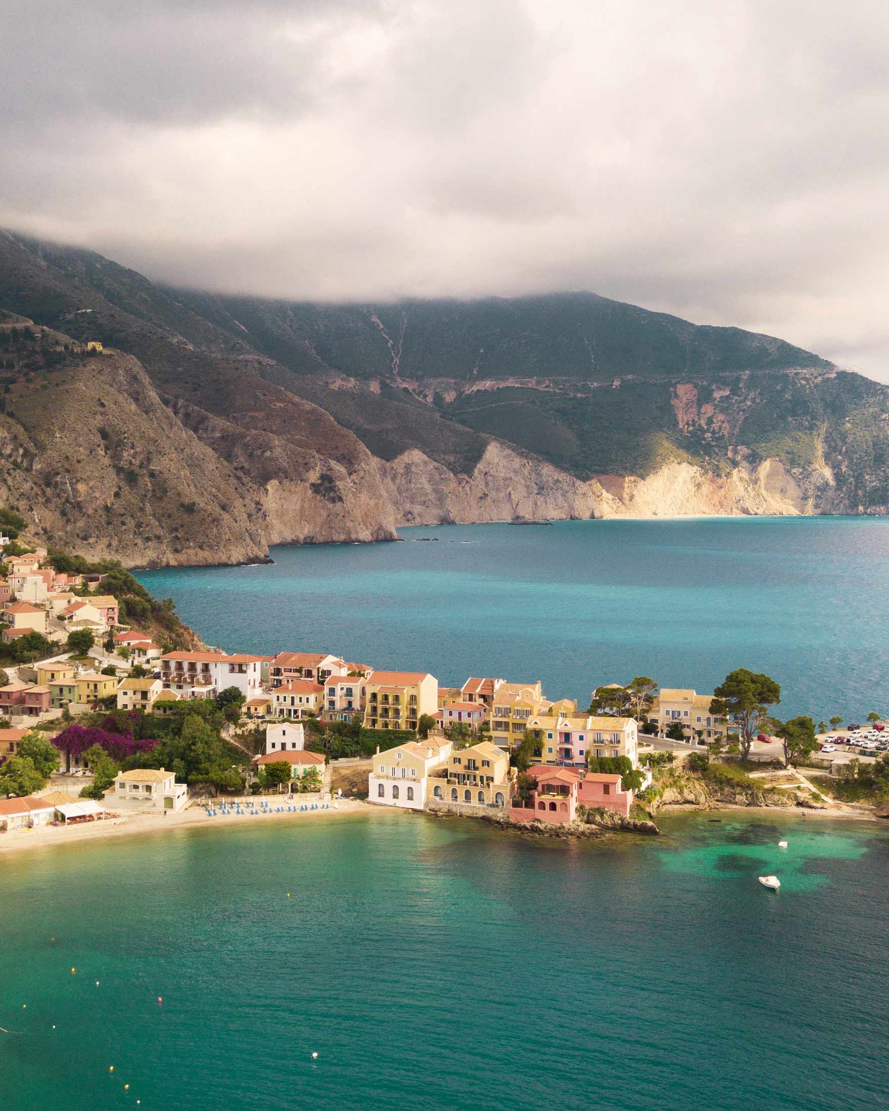 Eine Reihe farbenfroher Häuser an der Küste von Kefalonia, umgeben von türkisfarbenem Wasser und einer Bergwand im Hintergrund.