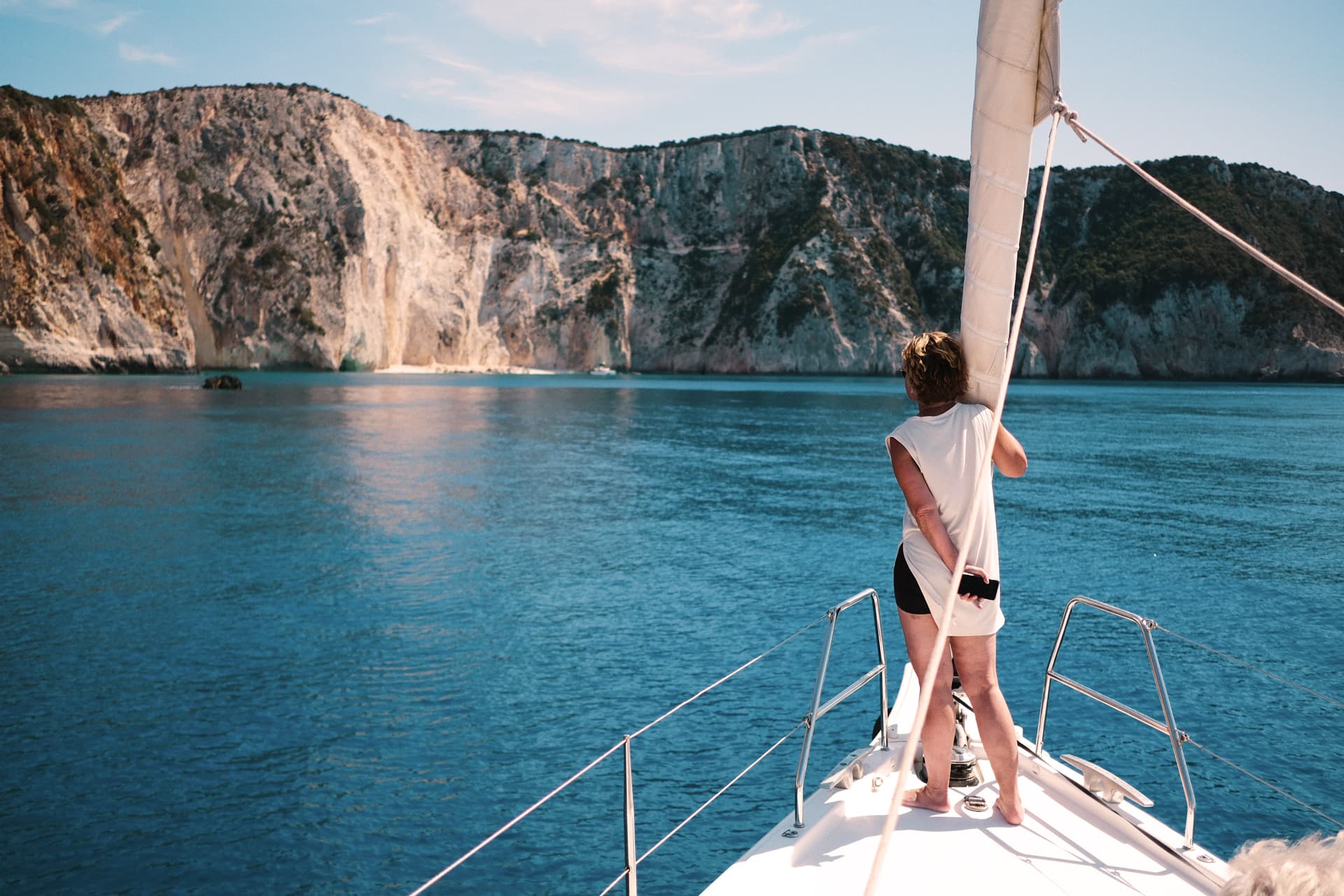 En kvinna står längst fram i fören på en segelbåt och tittar ut över höga klippor.