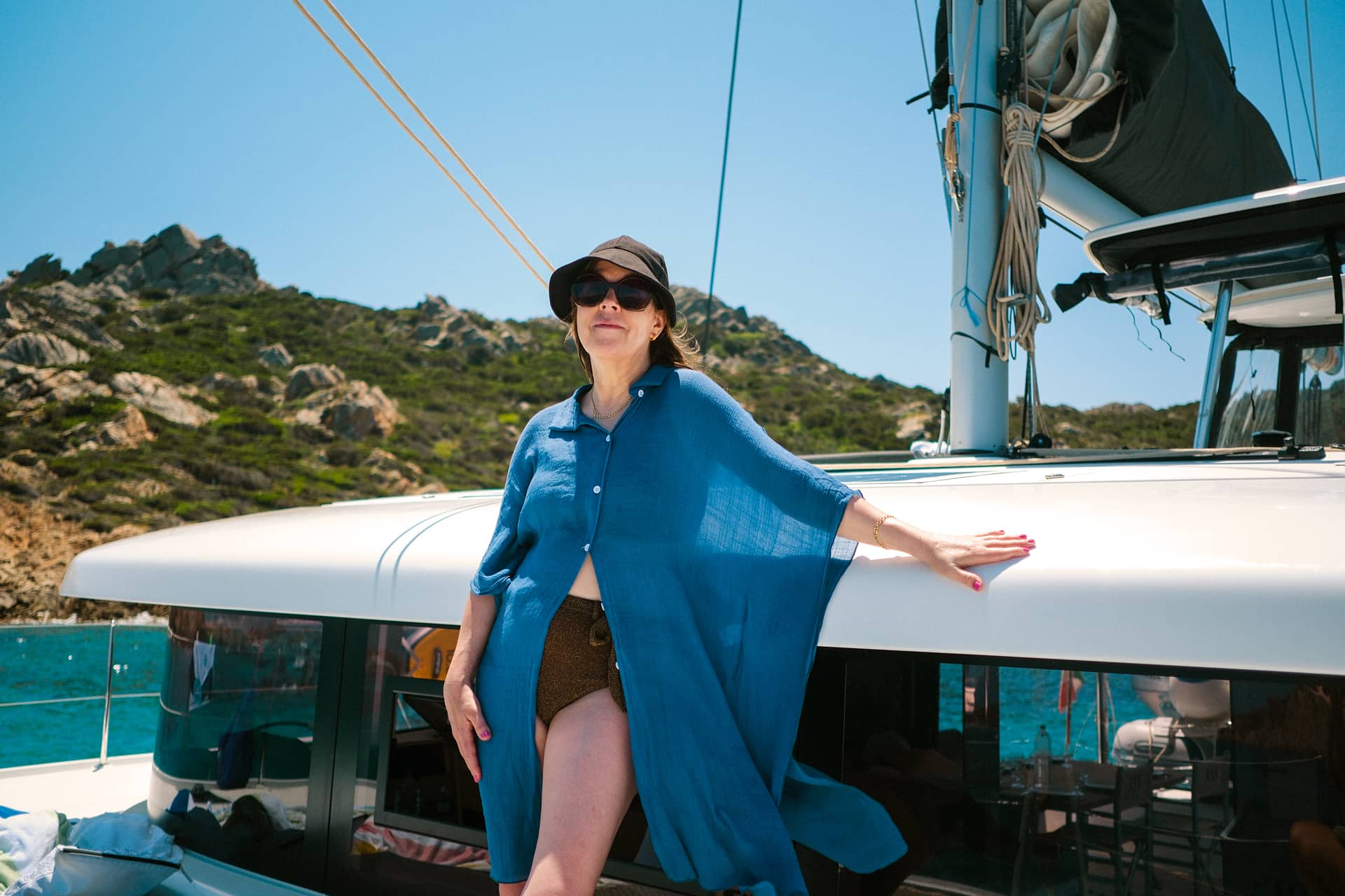 Woman standing on the foredeck wearing an indigo kaftan, hat and sunglasses.