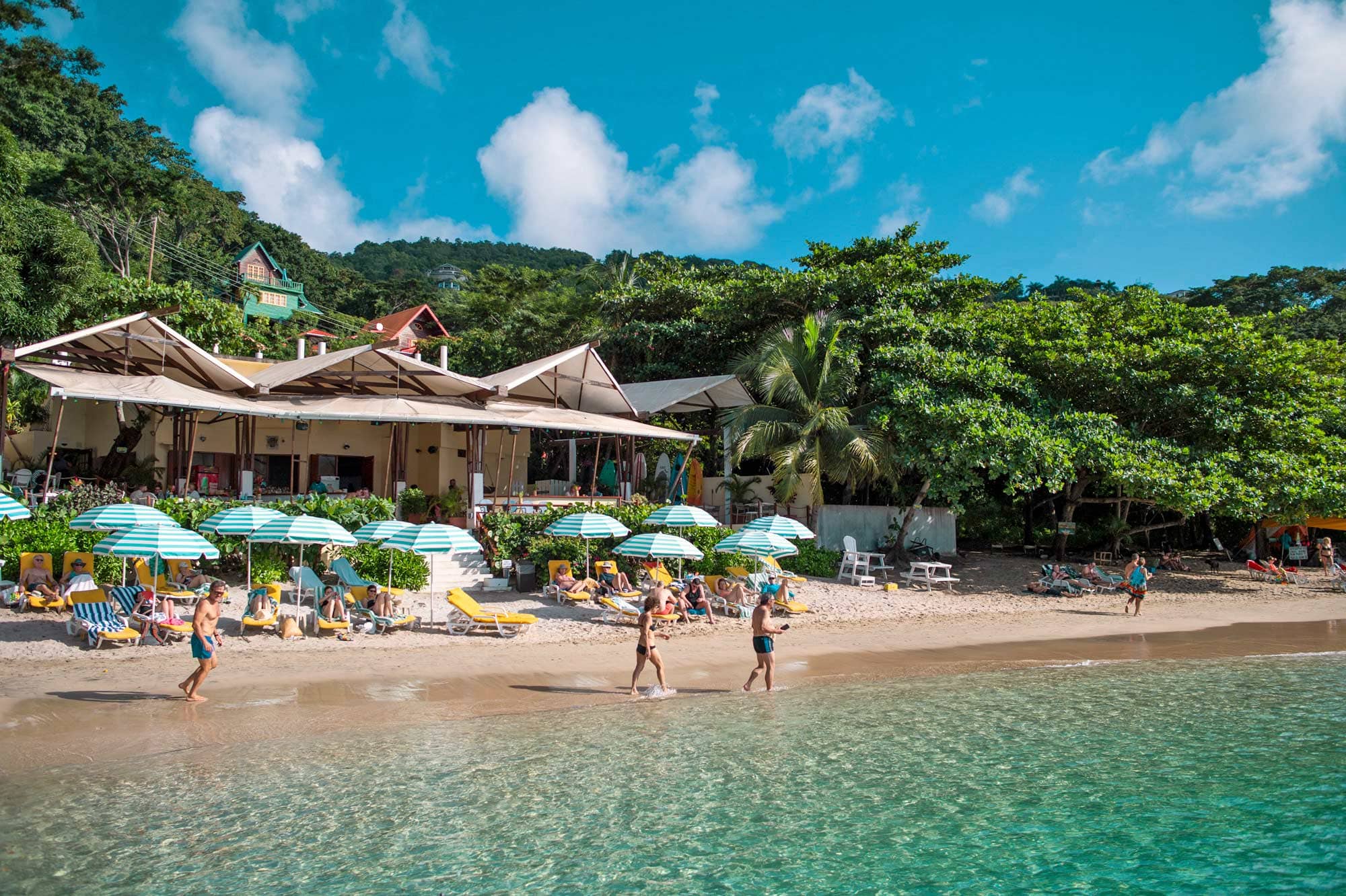 Strand vor einem Restaurant, 8 gestreifte Sonnenschirme und zwei beim Spazierengehen am Wasser.
