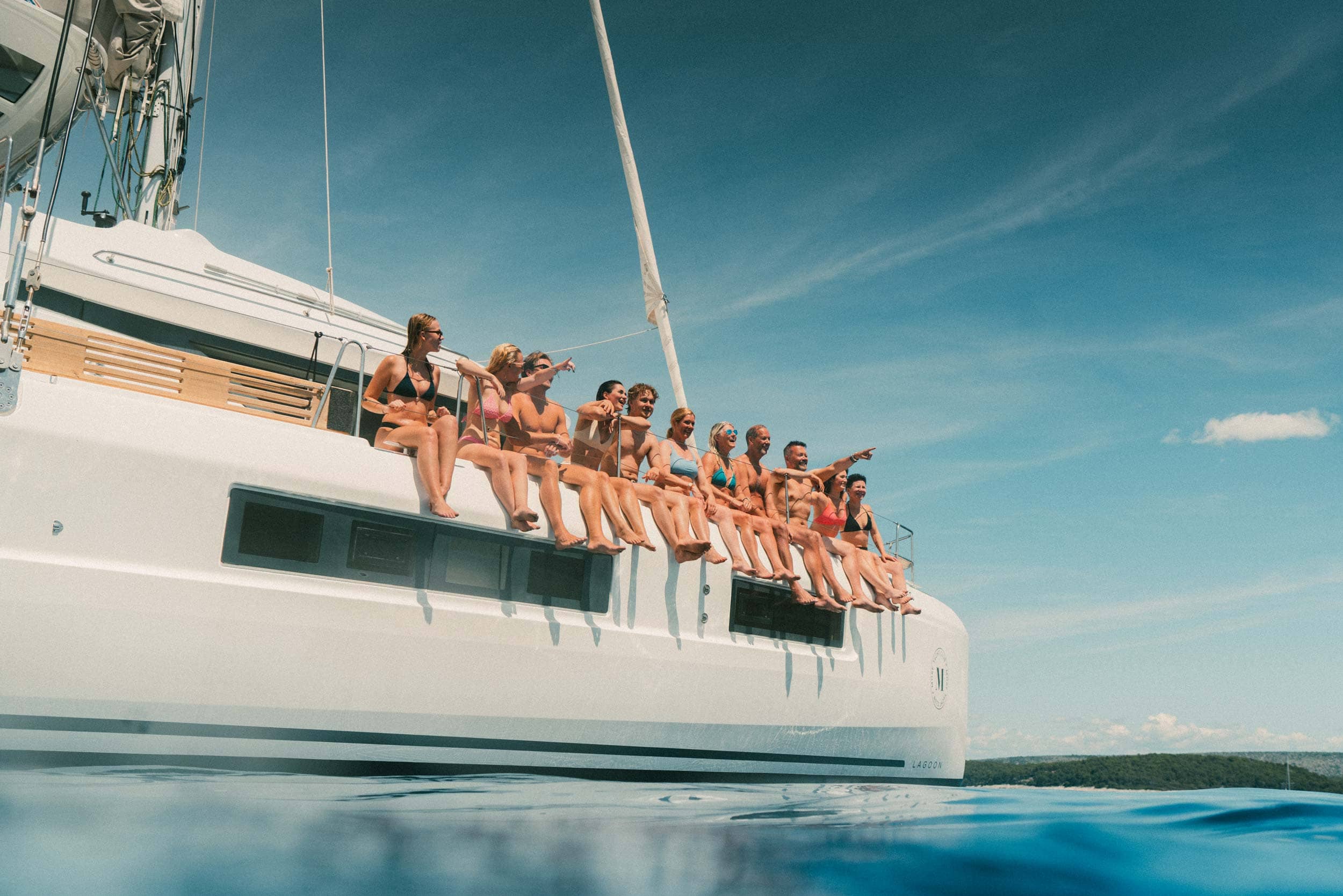 Eleven guests sit on the edge of a catamaran with their hands in the air